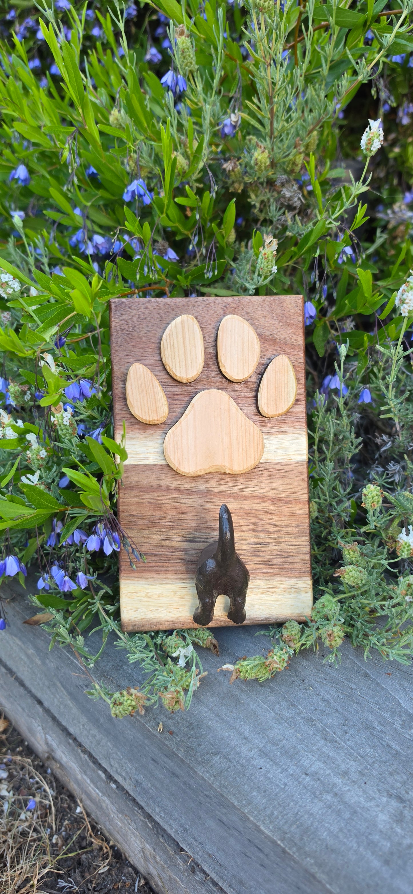 A rectangular wooden plaque or tray with a paw print design made from five natural wood pieces, featuring a small dark metal keyhole on the right side, photographed outdoors among green plants and small blue flowers next to a gray wooden surface.