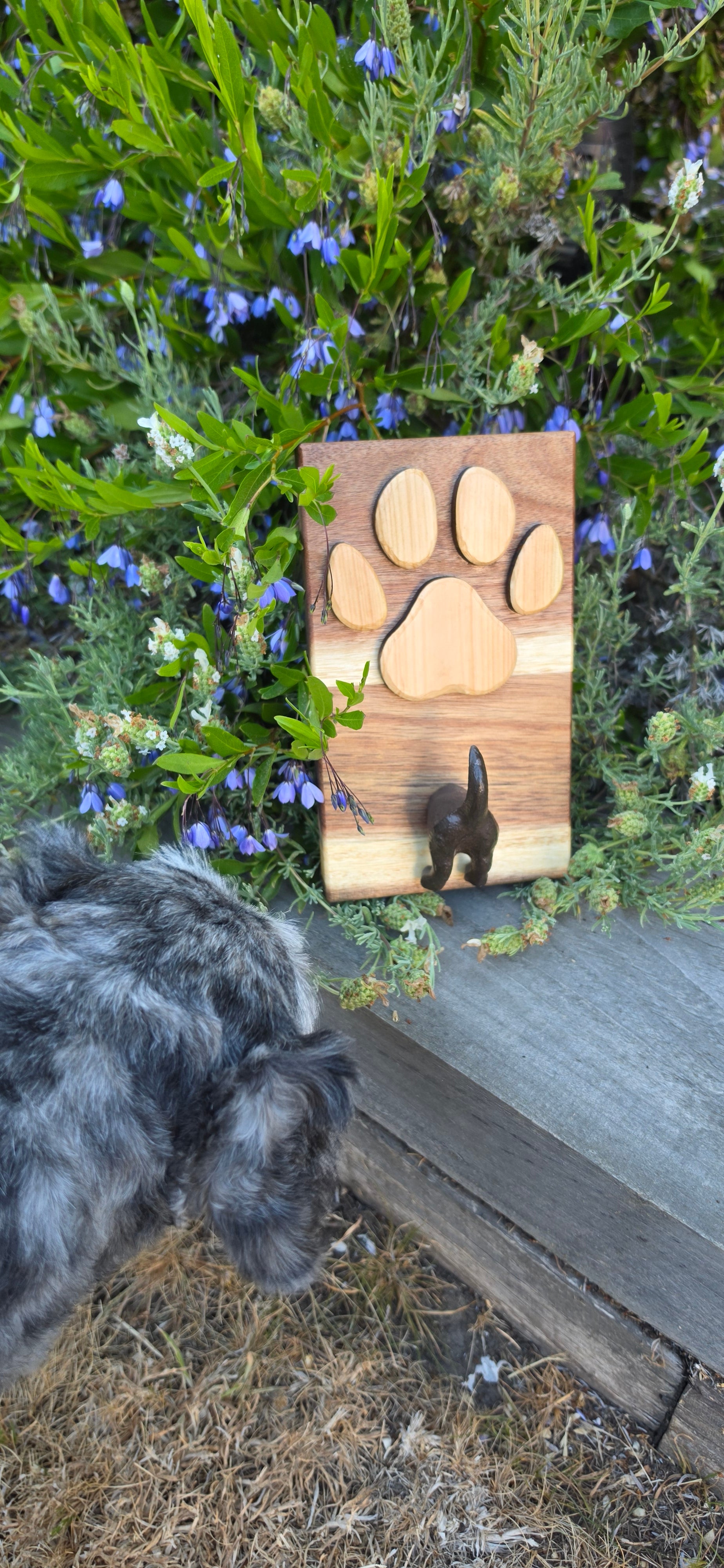 A small dog is lying on dry grass next to a wooden cutting board outdoors. The cutting board has four paw-print-shaped wooden pieces and a small black dog figurine.