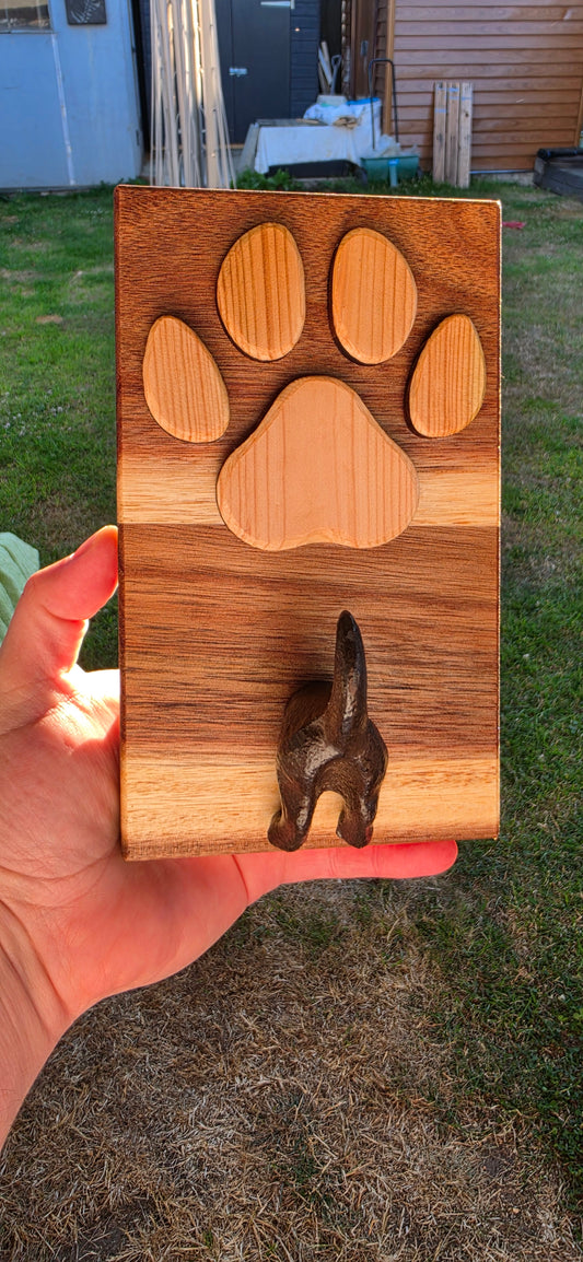 A hand holding a rectangular wooden box outdoors. The box has a carved paw print design in light wood and a dark metal dog-shaped hook on the right side.