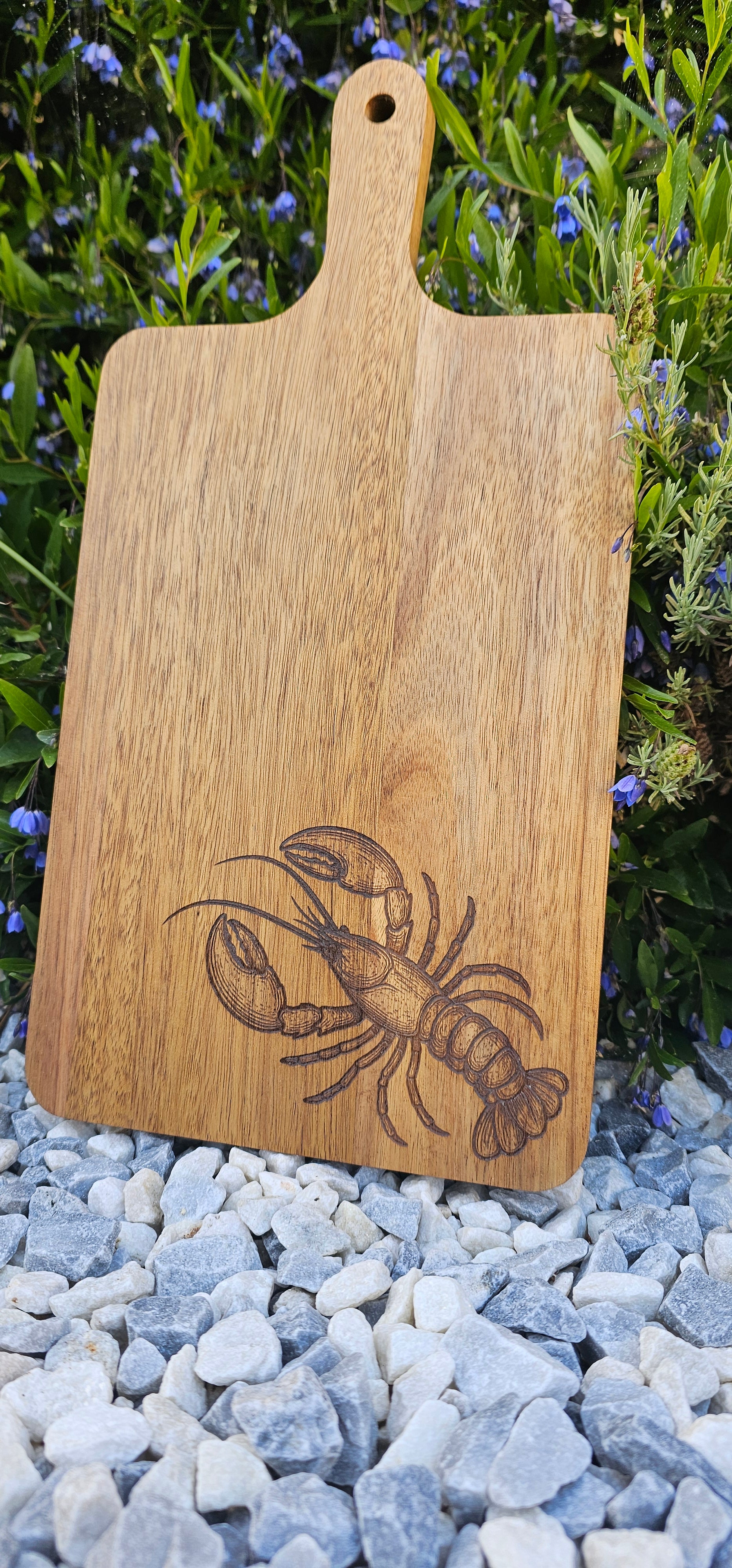 Wooden cutting board with a lobster engraving on a natural background of grass and stones.