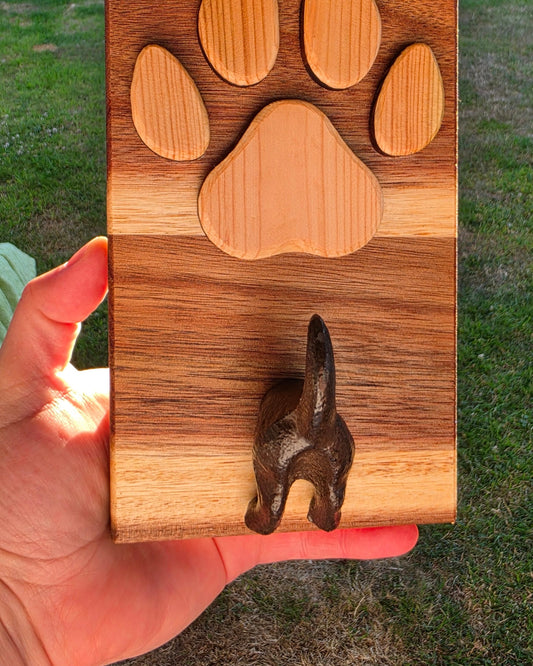 A hand holding a rectangular wooden box outdoors. The box has a carved paw print design in light wood and a dark metal dog-shaped hook on the right side.