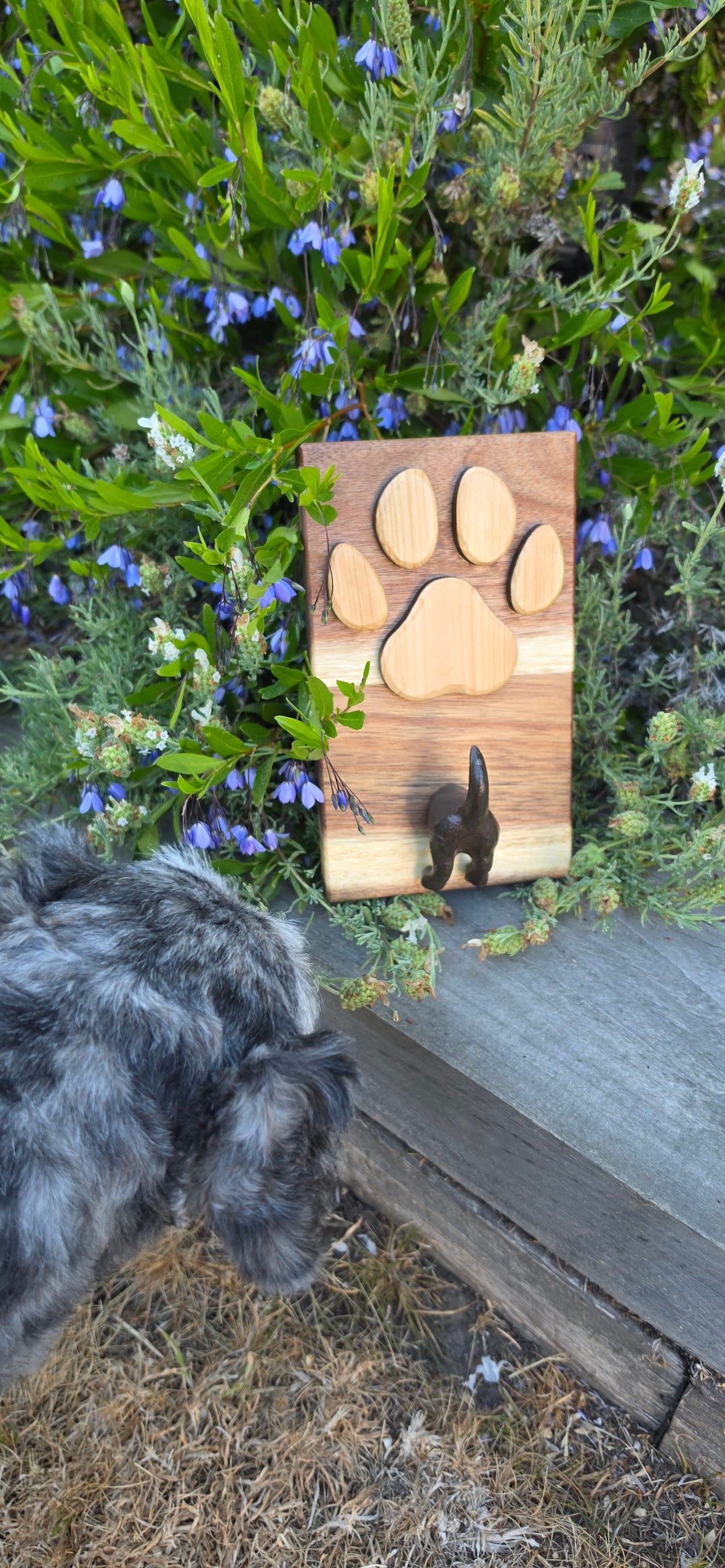A small dog is lying on dry grass next to a wooden cutting board outdoors. The cutting board has four paw-print-shaped wooden pieces and a small black dog figurine.