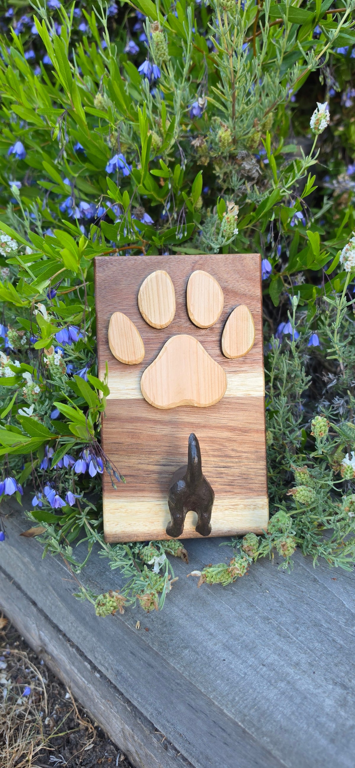 A rectangular wooden plaque or tray with a paw print design made from five natural wood pieces, featuring a small dark metal keyhole on the right side, photographed outdoors among green plants and small blue flowers next to a gray wooden surface.