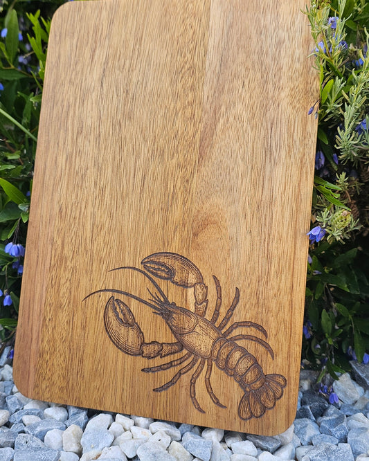 Wooden cutting board with a lobster engraving on a natural background of grass and stones.